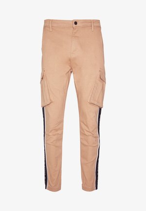 Beige cargobroek met zijzakken, voorzien van een taps toelopende pasvorm en een zwarte streep langs de zijkanten met witte tekstdetails. Gemaakt van een katoenmix.