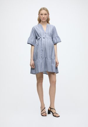 Terhes nő, kék csíkos, térdig érő ruhában, bővített ujjakkal és fekete magassarkú szandálban, egyszínű háttér előtt állva.