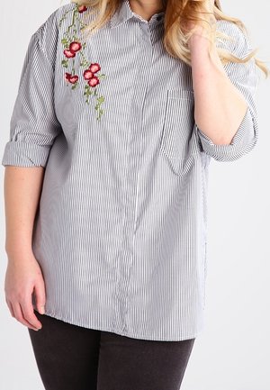 Kobieta nosząca czarno-białą koszulę w paski z długim rękawem podwiniętym na rękawach oraz czerwoną kwiatową haftowaną aplikacją na lewym ramieniu.