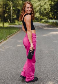 Pink wide-leg pants and a black cropped tank top, paired with black shoes. Holding a black wallet with polka dot accents. Outdoor setting.