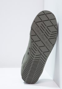 Grön canvas sneakers med en texturerad gummisula, som har ett hexagonalt mönster och synlig logotyp, vinklad för att visa undersidan.