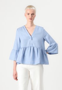 Lyseblå, langermet bluse med V-hals, knappdetaljer og volangkant, laget av teksturert stoff, kombinert med hvite bukser.