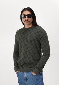 Olivgrüner, gestrickter Pullover mit strukturiertem Muster, rundem Halsausschnitt und gerippten Bündchen und Saum. Getragen mit blauen Jeans.