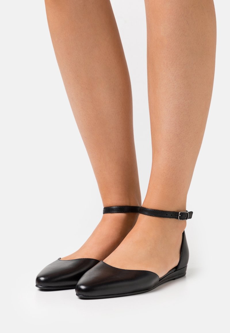 Chaussures plates en cuir noir avec brides autour de la cheville, portées pieds nus sur un fond blanc uni.