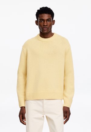 Joven con cabello corto y rizado, vestido con un suéter de punto amarillo claro y pantalones color marfil, de pie contra un fondo blanco.
