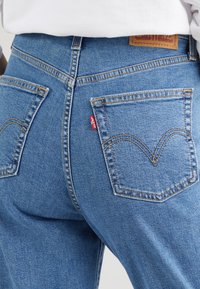 Närbild av blå Levi's jeans bakfickor med tan-färgade sömmar och en brun läderlogotyp på en person som bär en vit topp.