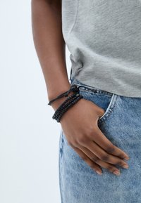 Juodas karoliukų apyrankė, sujungta su tekstūruotais siūlais, dėvima ant riešo. Kasdienių pilkų marškinėlių ir mėlynų džinsų derinys puikiai papildo aksesuarą.