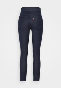 Jeans skinny de denim azul escuro com um cós alto, dois bolsos traseiros e uma pequena etiqueta de logotipo no cós direito. Textura suave.
