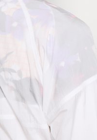 Giacca bianca leggera e semi-trasparente con dettaglio a stampa floreale. Tessuto strutturato e cuciture evidenziate, che mostrano un design a strati.