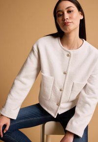 Weiße Wollmischungjacke mit rundem Halsausschnitt, zwei Fronttaschen und goldenen Knöpfen. Getragen mit blauer Denim, die lange Ärmel und einen lässigen Schnitt aufweist.