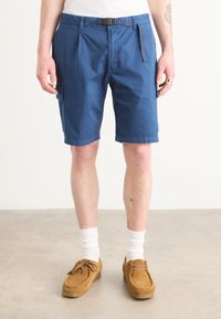 Shorts bleus en tissu mélangé coton avec un devant plat, des poches latérales et une boucle de ceinture. Associés à des mocassins beiges et des chaussettes blanches.