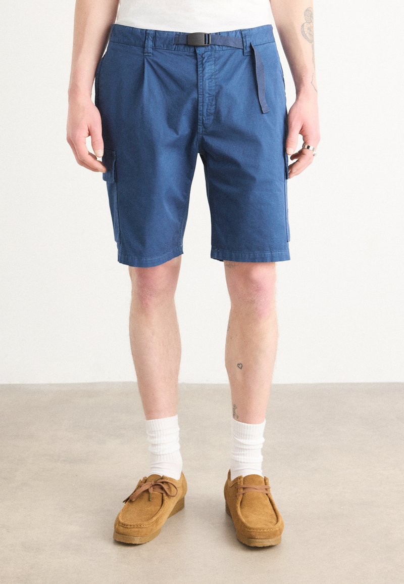 Shorts bleus en tissu mélangé coton avec un devant plat, des poches latérales et une boucle de ceinture. Associés à des mocassins beiges et des chaussettes blanches.