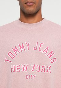 Rozā džemperis ar apaļu kakla izgriezumu, kurā ar lielajiem baltajiem un rozā burtiem ir uzraksts "TOMMY JEANS", bet zem tā mazākajā tekstā "NEW YORK CITY".