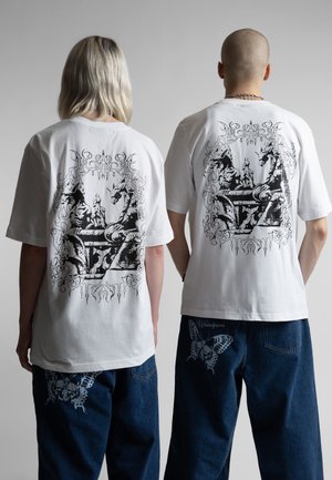 To personer iført hvide t-shirts med grafiske tryk af en sort drage og et bord på ryggen, kombineret med mørkeblå jeans med sommerfugleprint.