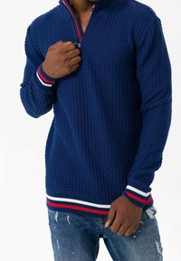 Maglione blu navy con zip e motivo testurizzato, con accenti a strisce rosse e bianche sul colletto e sui polsini. Indossato sopra jeans blu.