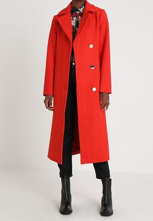 Persona che indossa un lungo cappotto rosso acceso con tre bottoni, pantaloni neri, stivali neri e una camicia con motivo floreale sotto.