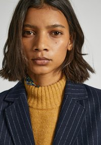 Blazer azul marino a rayas con solapas acanaladas, combinado con un suéter mostaza de canalé. Acento notable de tela floral en el cuello. Cabello corto y oscuro.