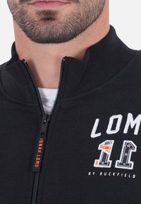 Veste zippée noire avec col montant, texte blanc brodé et étiquette orange avec "JONAH LOMU." Tissu lisse au design sportif.