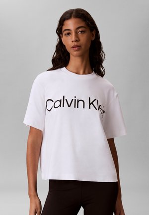 Jeune femme aux longs cheveux foncés portant un t-shirt blanc ample avec le texte « Calvin Klein » et un pantalon noir, debout devant un fond gris uni.