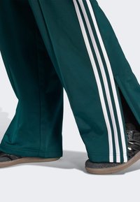 Teal athletic pants with three white stripes down the sides, featuring side slits at the hem. Paired with black sports shoes.
