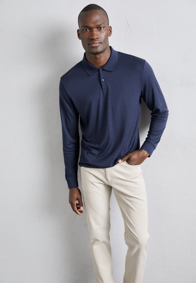 Marineblaues Langarm-Polohemd mit Kragen und Knopfdetails, kombiniert mit hellbeigen Slim-Fit-Hosen. Glatte Stoffstruktur, lässige Passform.