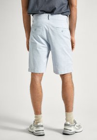 Uomo che indossa pantaloni corti blu chiaro al ginocchio, calze beige e sneakers grigie e bianche, in piedi contro uno sfondo chiaro e semplice.