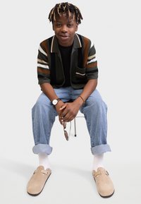 Sweater in green with orange and white stripes, black t-shirt, light blue jeans, beige clogs, white socks, holding sunglasses, seated on a chair.