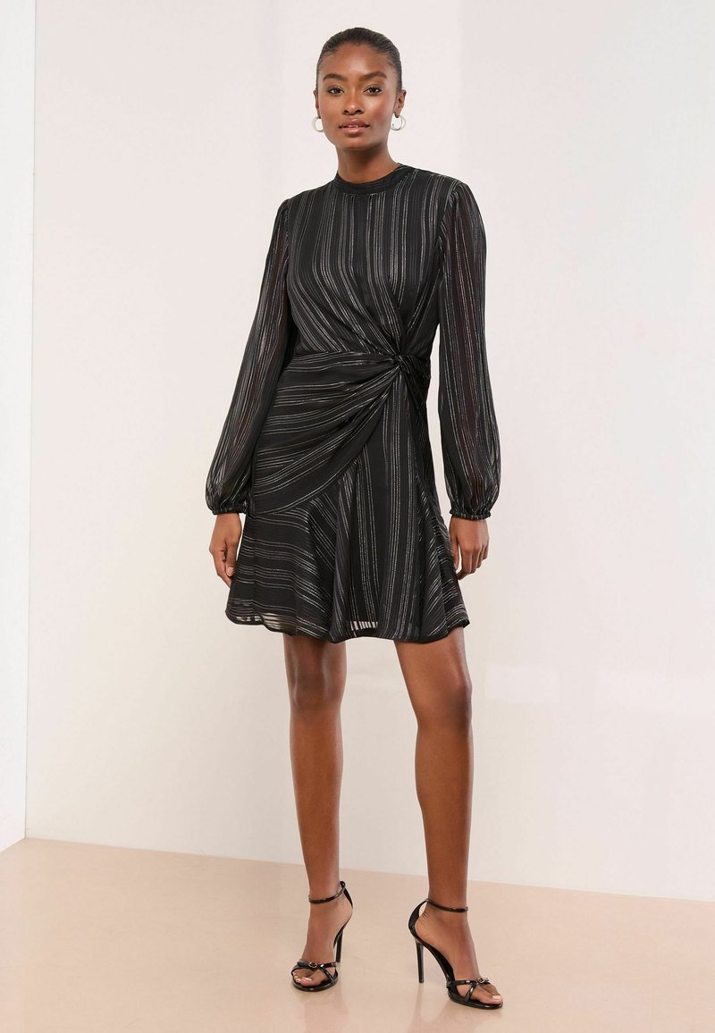 Černé šaty s metalickými stříbrnými pruhy, dlouhými rukávy, s twistem v pase a rozšířenou sukní, doplněné černými sandály na vysokém podpatku.