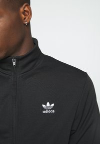 Giacca nera con zip realizzata in tessuto testurizzato, con un piccolo logo Adidas bianco sul petto e un design con colletto alto.