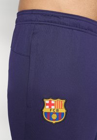Paarse sportshorts van een textuurstof, met een elastische tailleband en een geborduurd FC Barcelona-logo aan de zijkant.