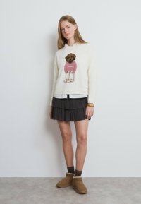 Cremefarbener Pullover mit einem gestrickten Bärenmotiv in Pink und Braun, kombiniert mit einem mehrlagigen schwarzen Rock und braunen Stiefeletten mit Fellbesatz.