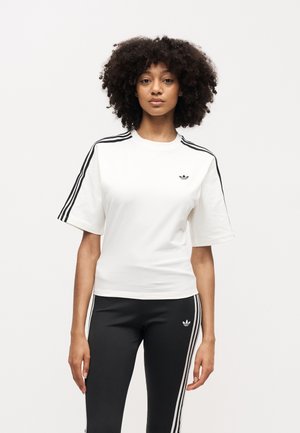 Kvinde med krøllet hår iført en hvid Adidas T-shirt med sorte striber på ærmerne og sorte Adidas leggings med hvide striber.