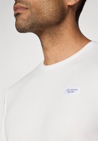 Biele tričko s krátkym rukávom v textúrovanej tkanine s malou modro-červenou nášivkou "Ted Baker Sport" na hornej ľavej strane.