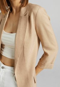 Blazer en lin beige avec une coupe décontractée, col cranté, manches trois-quarts retroussées, assorti à un crop top blanc et un jean clair.