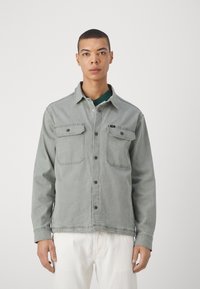 Chaqueta de mezclilla gris claro con dos bolsillos en el pecho, cierre de botones y un cuello. Llevada sobre una camisa verde oscura. Pantalones blancos.