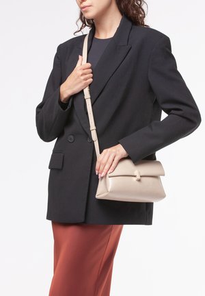 Beige leren crossbody tas met een klep, rechthoekige vorm en subtiele textuur. De schouderband is verstelbaar. Gecombineerd met een zwarte blazer en roestkleurige broek.