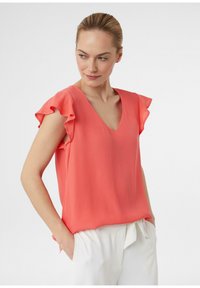 Korallefarbene Bluse mit kurzen Ärmeln und Rüschekanten, im V-Ausschnitt-Design, lockere Passform und glatter Stoff, kombiniert mit weißen maßgeschneiderten Hosen.
