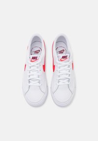 Zapatillas Nike blancas con logotipos de swoosh rojos y cordones blancos vistas desde arriba sobre un fondo blanco liso.