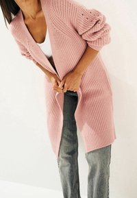 Cárdigan largo y abierto en punto rosa claro, con textura acanalada y mangas voluminosas. Combinado con jeans grises.