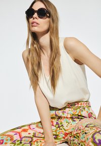Sunglasses med sköldpaddsdesign, rund form och mörka linser. Modell iförd en krämfärgad ärmlös topp och livfulla mönstrade byxor.