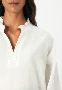 Hvit bluse med volangkrage, V-hals og dekorativ blondekant. Stoffet har små hevede prikker som gir tekstur.
