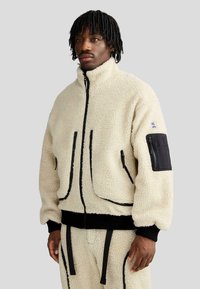 Giacca in fleece color crema con colletto alto, dettagli neri, tasche con cerniera e tessuto texturizzato, indossata con pantaloni coordinati.