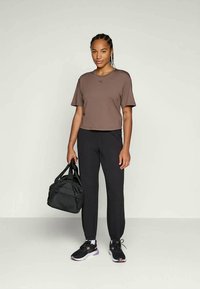Kvinde står iført en brun kortærmet skjorte, sorte joggers, sorte sneakers og holder en sort træningstaske i højre hånd imod en ensfarvet lys baggrund.