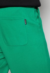 Grüne Baumwoll-Sweatpants mit glatter Textur, ausgestattet mit einer großen Gesäßtasche und einem schwarzen Markenetikett. Das Design umfasst einen elastischen Bund.