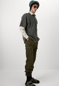 Chemise noire et grise à rayures oversize à manches courtes superposée sur un haut à manches longues blanc ; pantalon cargo vert olive ; chaussettes sombres avec des sandales.