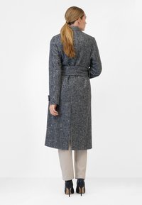Lång grå tweedkappa med strukturerad finish, bälte i midjan och slits bak i mitten. Matchad med beige byxor och svarta klackar.