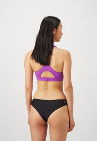 Sutiã esportivo roxo com design nas costas tipo "racerback" e detalhe recortado, combinado com calções de bikini pretos. Feito de material elástico e que absorve a humidade.