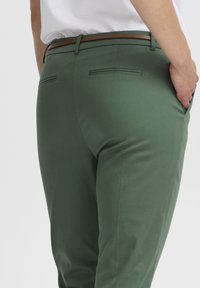Pantalons verts avec une texture lisse, dotés d'une ceinture marron, de poches latérales et de rabats bi-matière à l'arrière, ajustés pour une coupe près du corps.