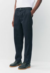 Dark blue denim trousers with a straight cut, featuring a single button closure and side pockets, paired with black footwear.