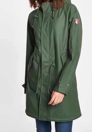 Imperméable vert à capuche avec fermeture éclair avant et cordons, porté par une personne aux cheveux bruns et jean bleu sur fond blanc.